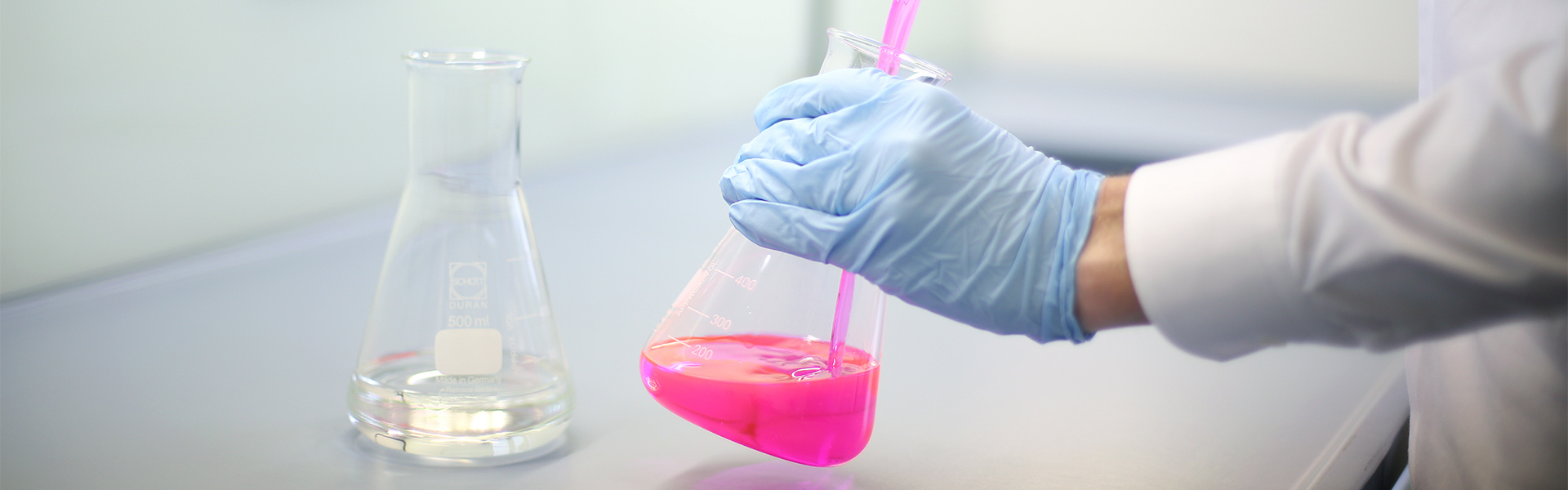Darstellung eines Labor-Mitarbeiters mit Gummihandschuhen, der eine pinke Flüssigkeit in einem Erlmeier-Kolben umrührt