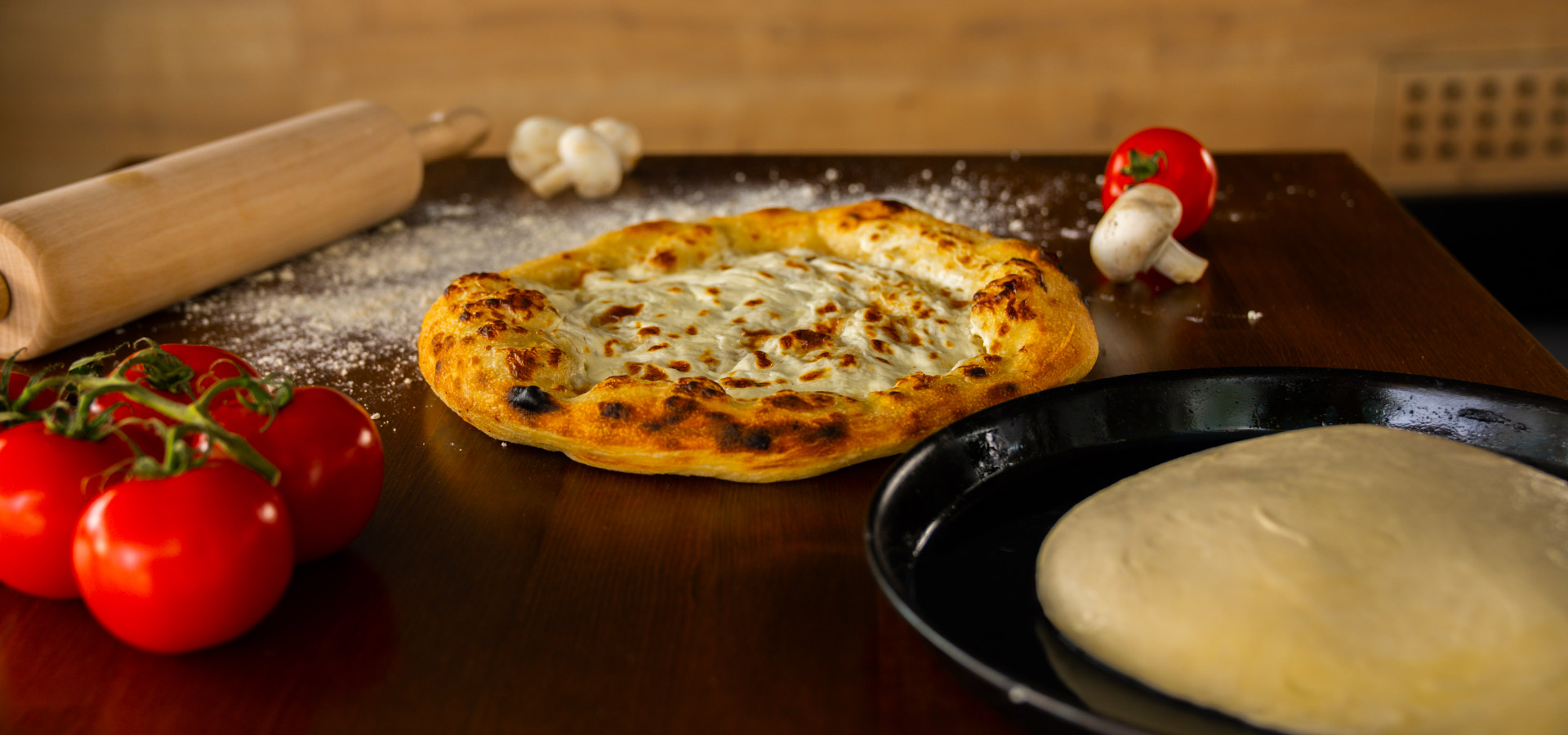 Eine gebackene Pizza liegt auf einem Holztisch, umgeben von Tomaten, Pilzen und einem Nudelholz