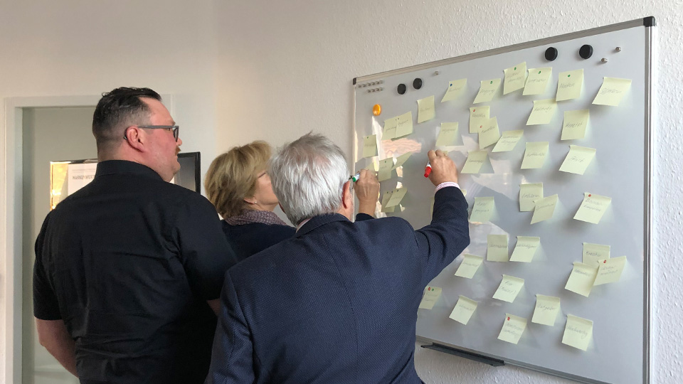 Workshopsituation in der drei Personen vor einem Whiteboard stehen, eine der Personen schreibt auf eines der Post-Its