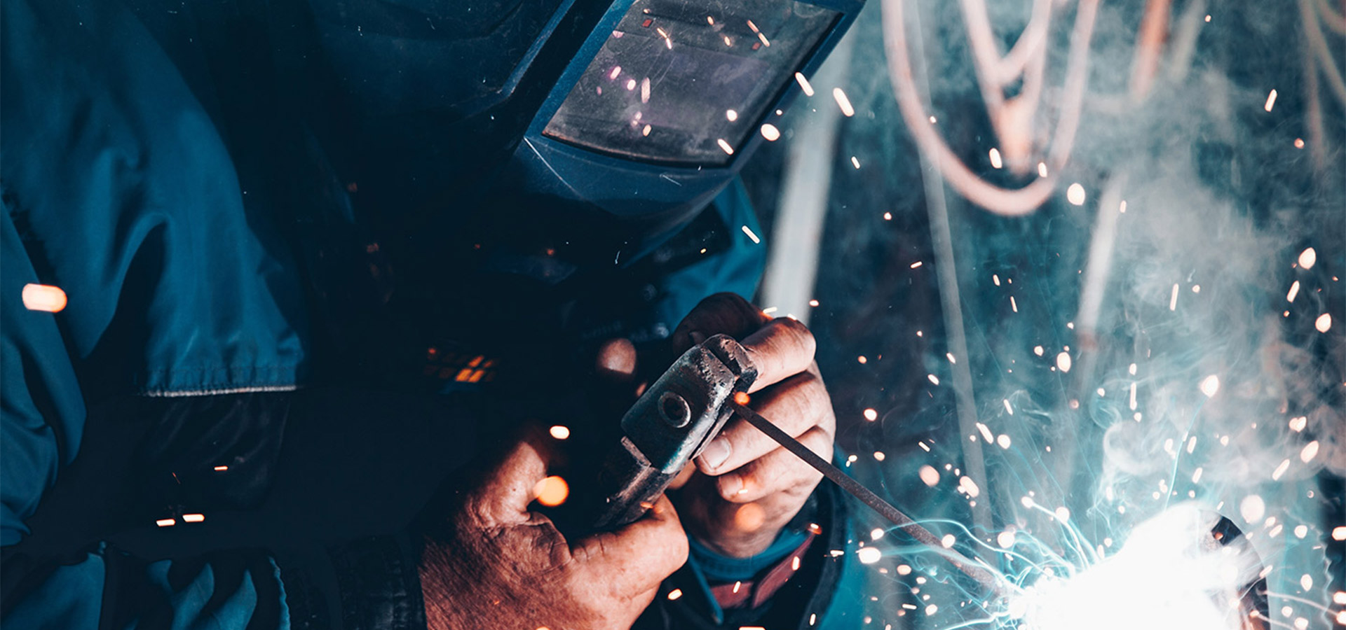 Schweißer in Schutzkleidung schweißt mit einem Schweißbrenner ein Metallstück an, helle Funken spühen