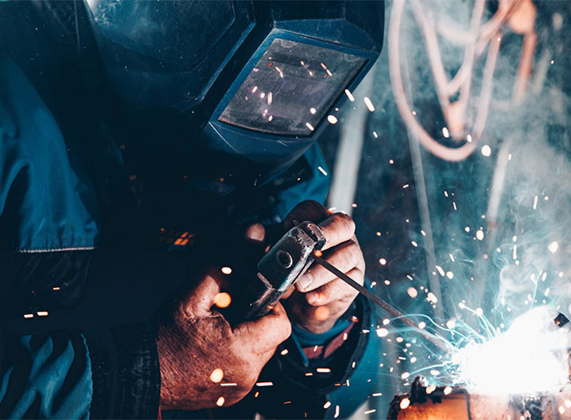 Schweißer in Schutzkleidung schweißt mit einem Schweißbrenner ein Metallstück an, helle Funken spühen