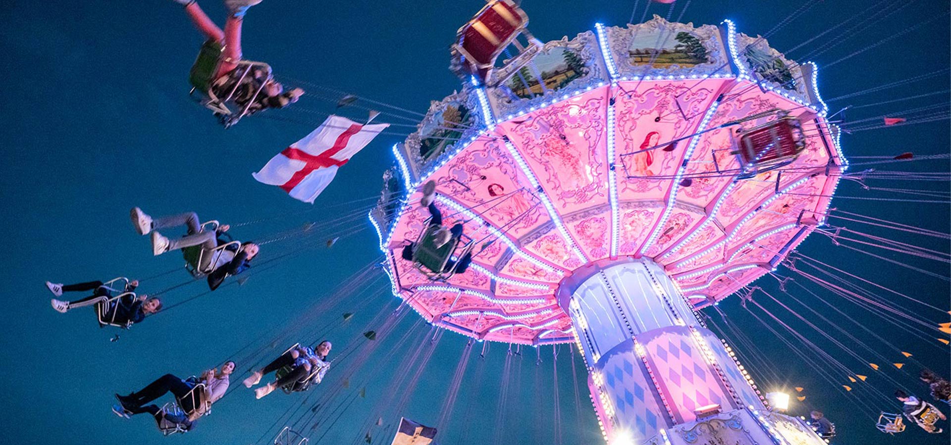Pink angestrahltes Kettenkarussell im Betrieb, auf dem sieben Gäste fahren und an dem eine englische Flagge hängt
