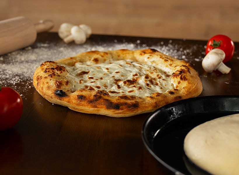 Eine gebackene Pizza liegt auf einem Holztisch, umgeben von Tomaten, Pilzen und einem Nudelholz